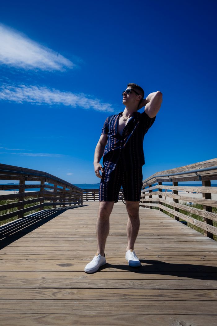 Man in sunglasses striking a pose on a wooden boardwalk under a bright blue sky.